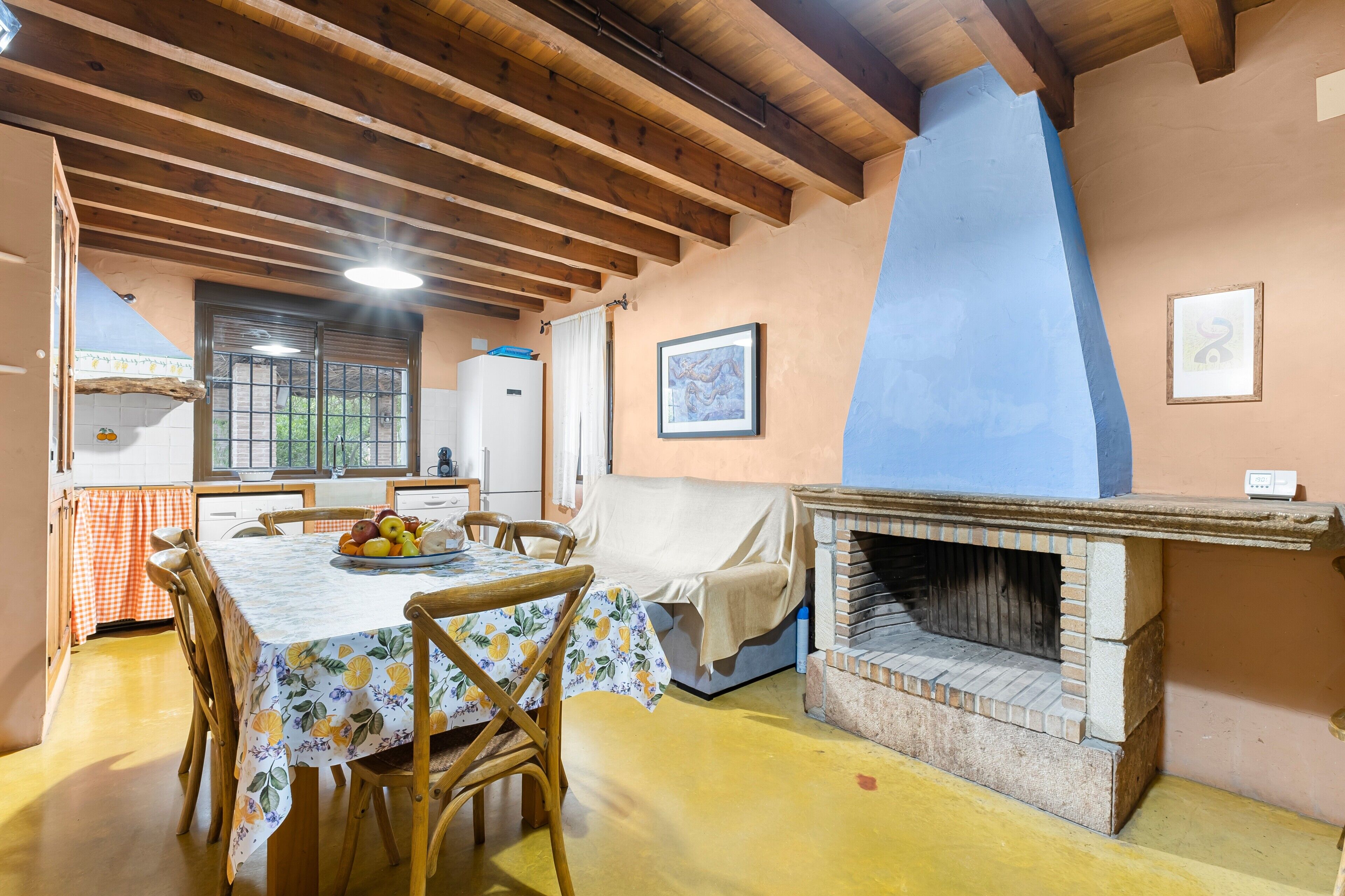 A dining area with a table set for four, wooden chairs, a pizza oven, and a window with curtains.