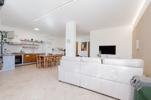 Une cuisine moderne avec des meubles en bois, un coin repas avec des chaises, un canapé blanc et un téléviseur à écran plat fixé au mur.