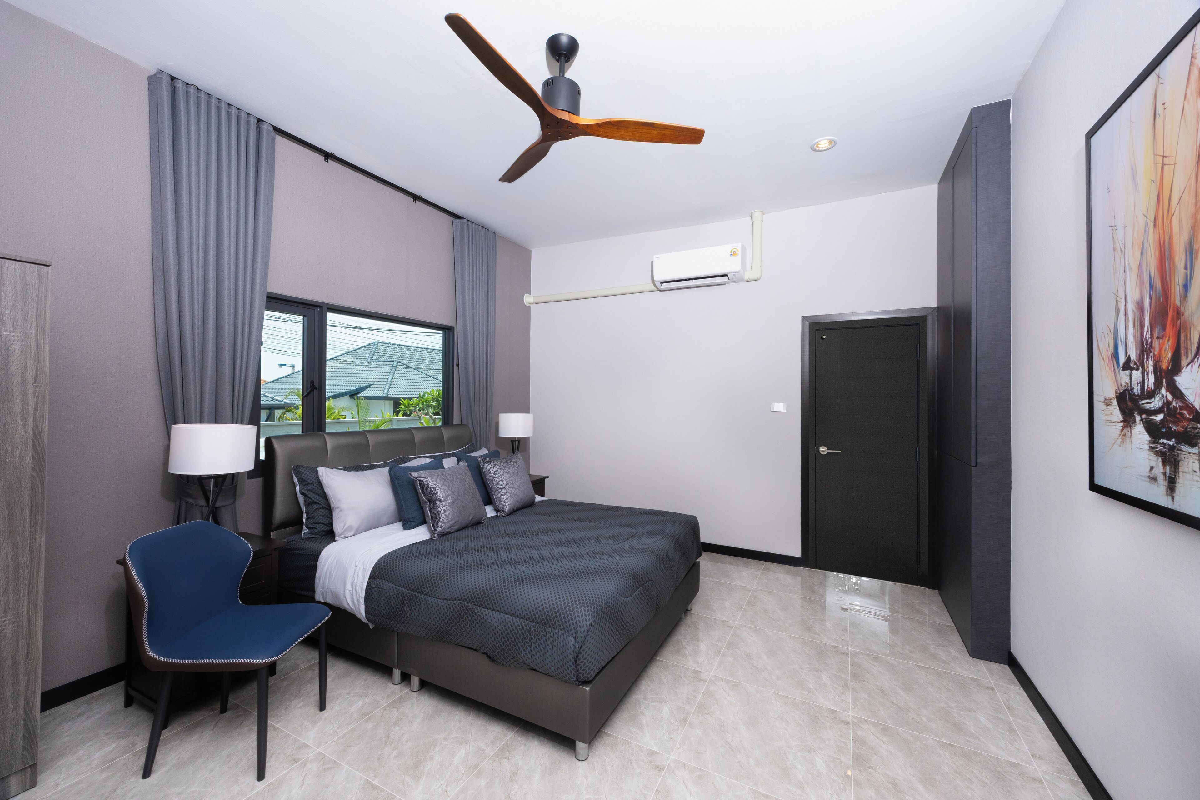 A modern hotel room with a bed, a chair, a ceiling fan, an air conditioning unit, and a door.