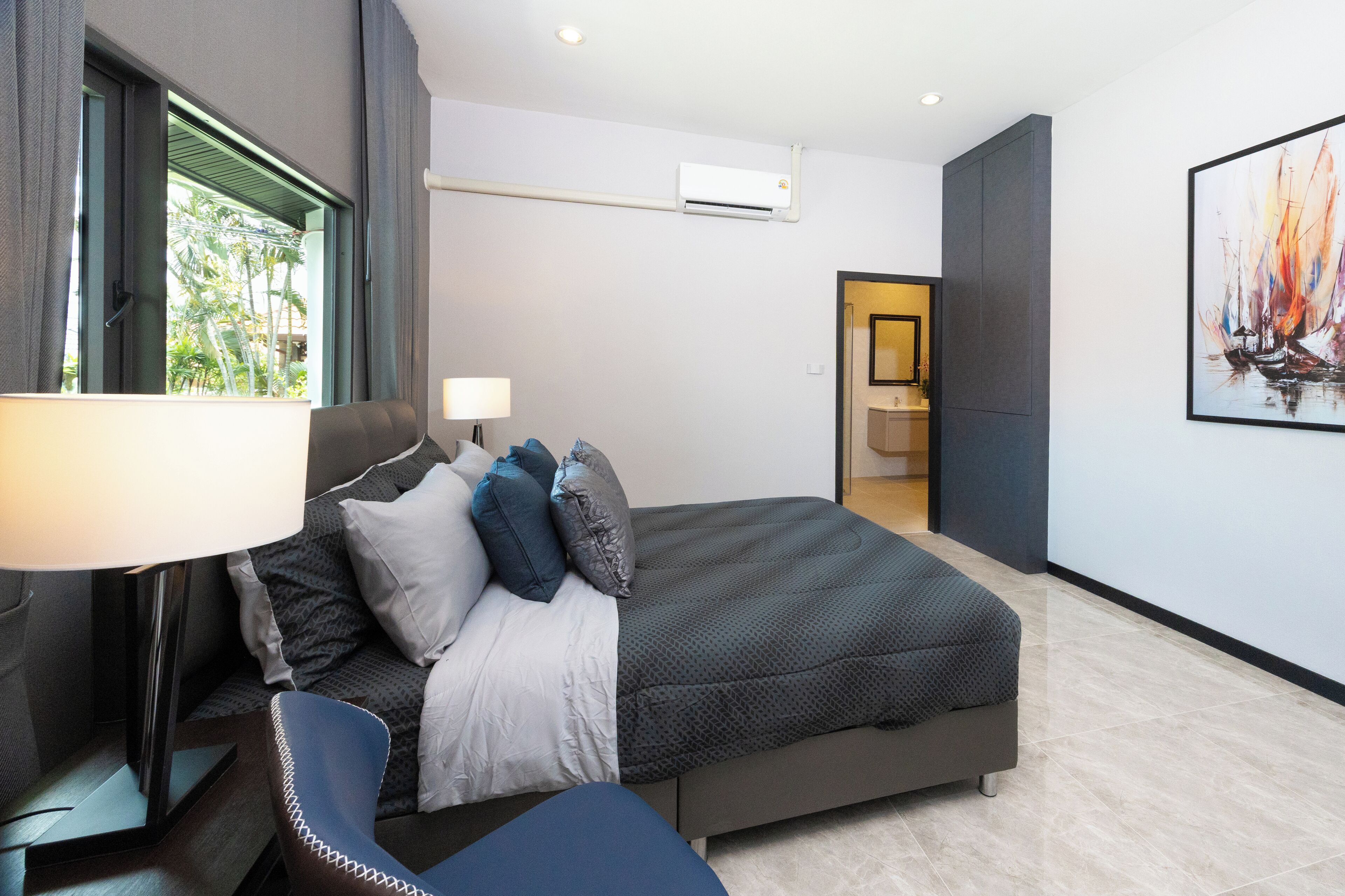 Quarto de hotel moderno com uma cama grande, uma escrivaninha com abajur, uma cadeira e um quadro na parede.