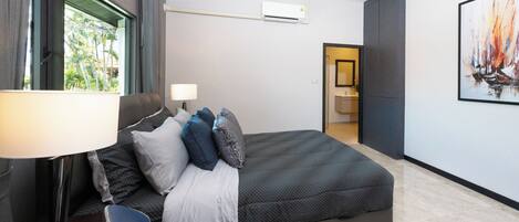 Quarto de hotel moderno com uma cama grande, uma escrivaninha com abajur, uma cadeira e um quadro na parede.