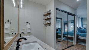 Un baño moderno con un espejo grande, un lavamanos y una ducha. Hay un estante de madera con artículos de aseo y una puerta corredera de cristal que da a un dormitorio con cama y ventana.