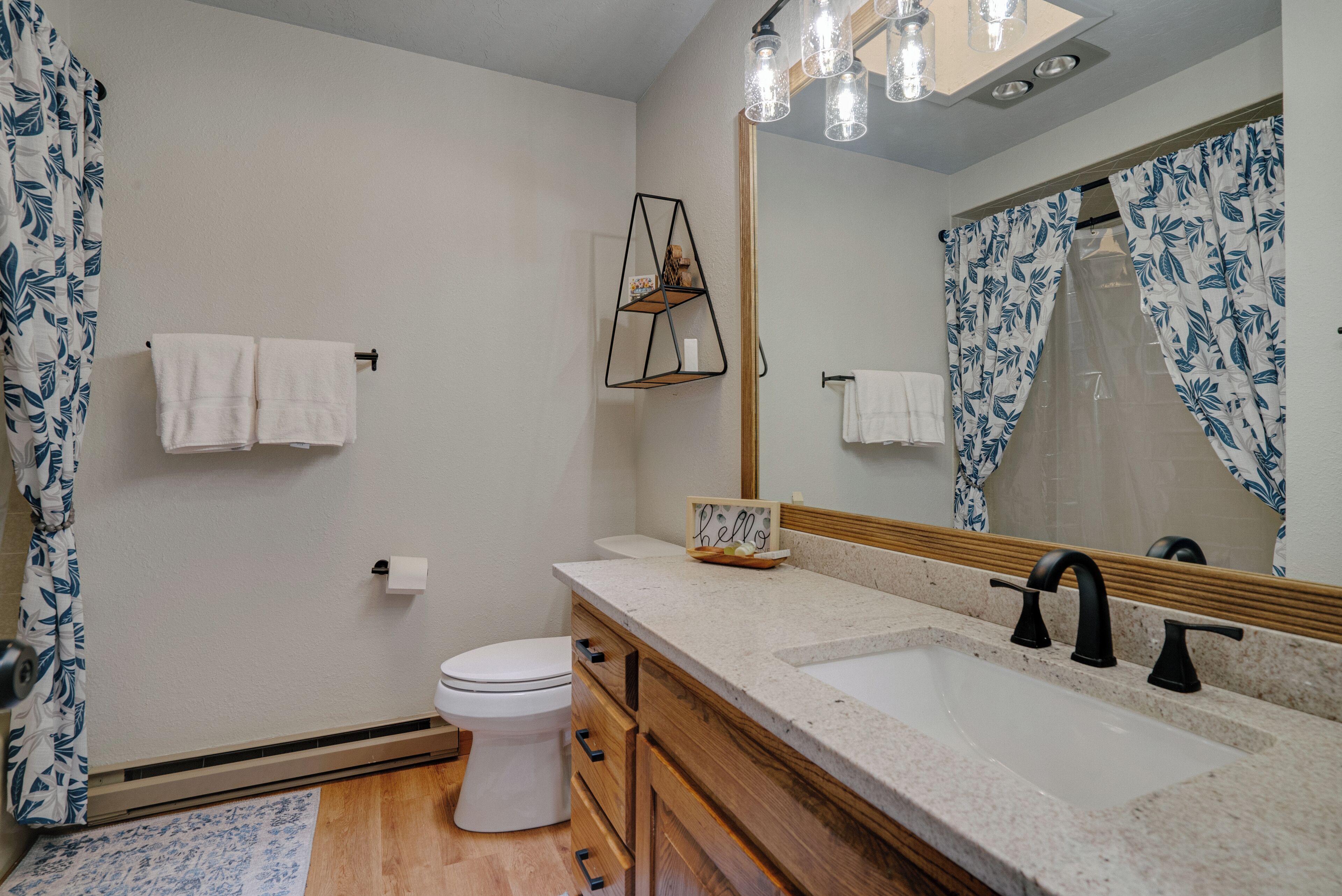 A bathroom with a white toilet, wooden cabinets, a sink with a black faucet, a mirror, and a shower curtain with a leaf pattern.