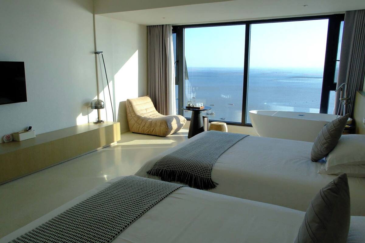 A modern hotel room with two beds, a chair, a small table, and a large window offering a sea view.