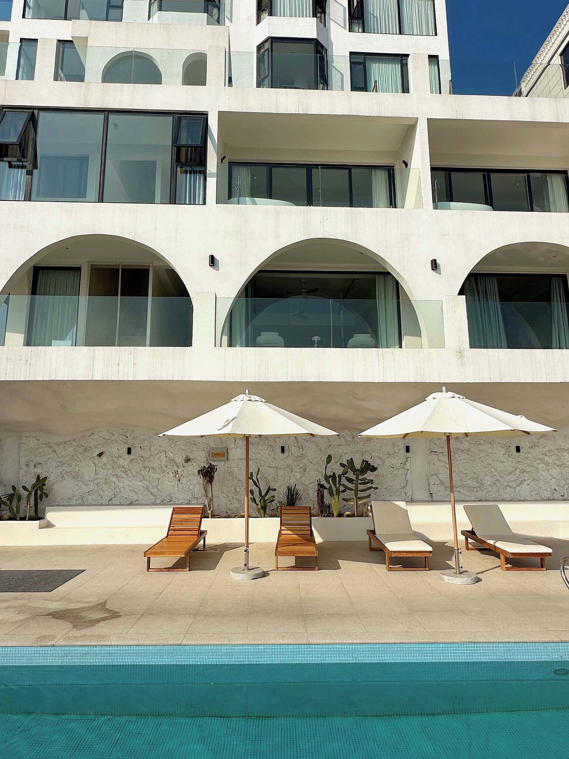 Un hotel moderno con zona de piscina que incluye tumbonas y sombrillas, fachada blanca y ventanas arqueadas.
