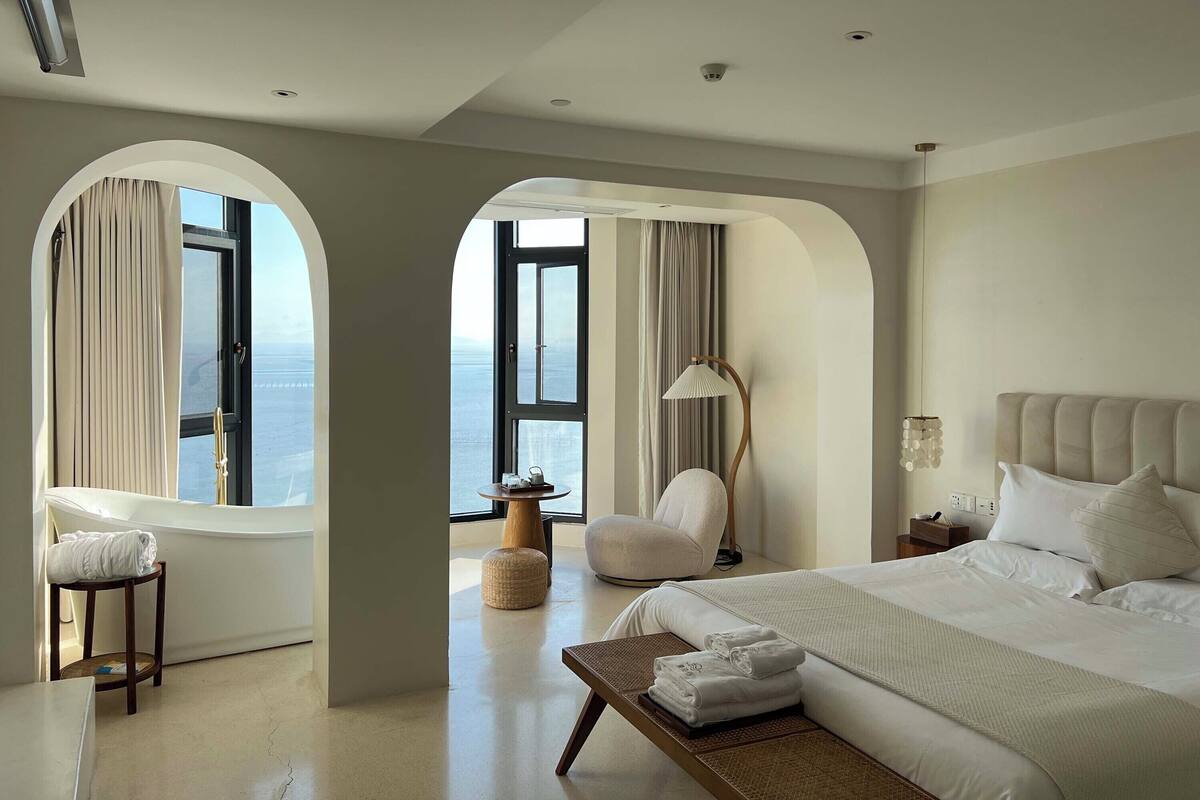 A modern hotel room with a large bed, a chair, a side table, and a view of the sea through arched windows.