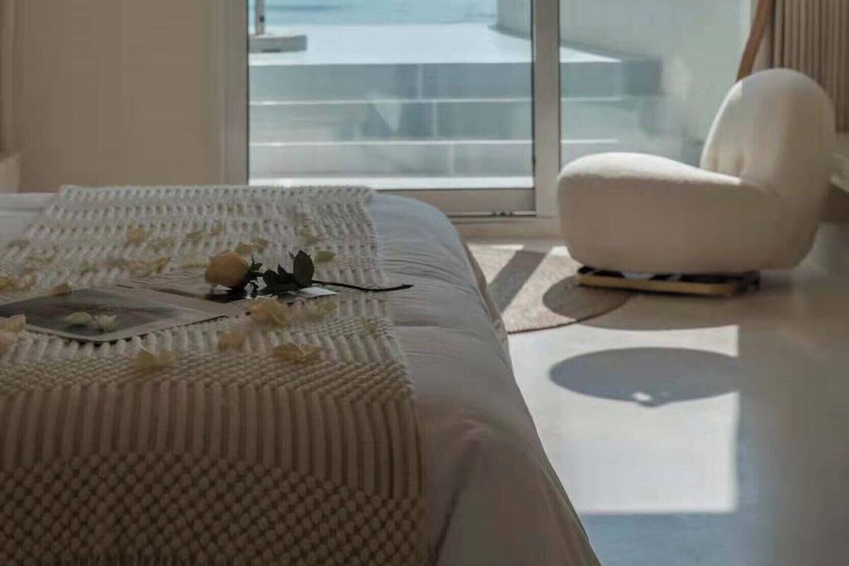 A modern hotel room with a large bed, a white armchair, and a view of the ocean through floor-to-ceiling windows.