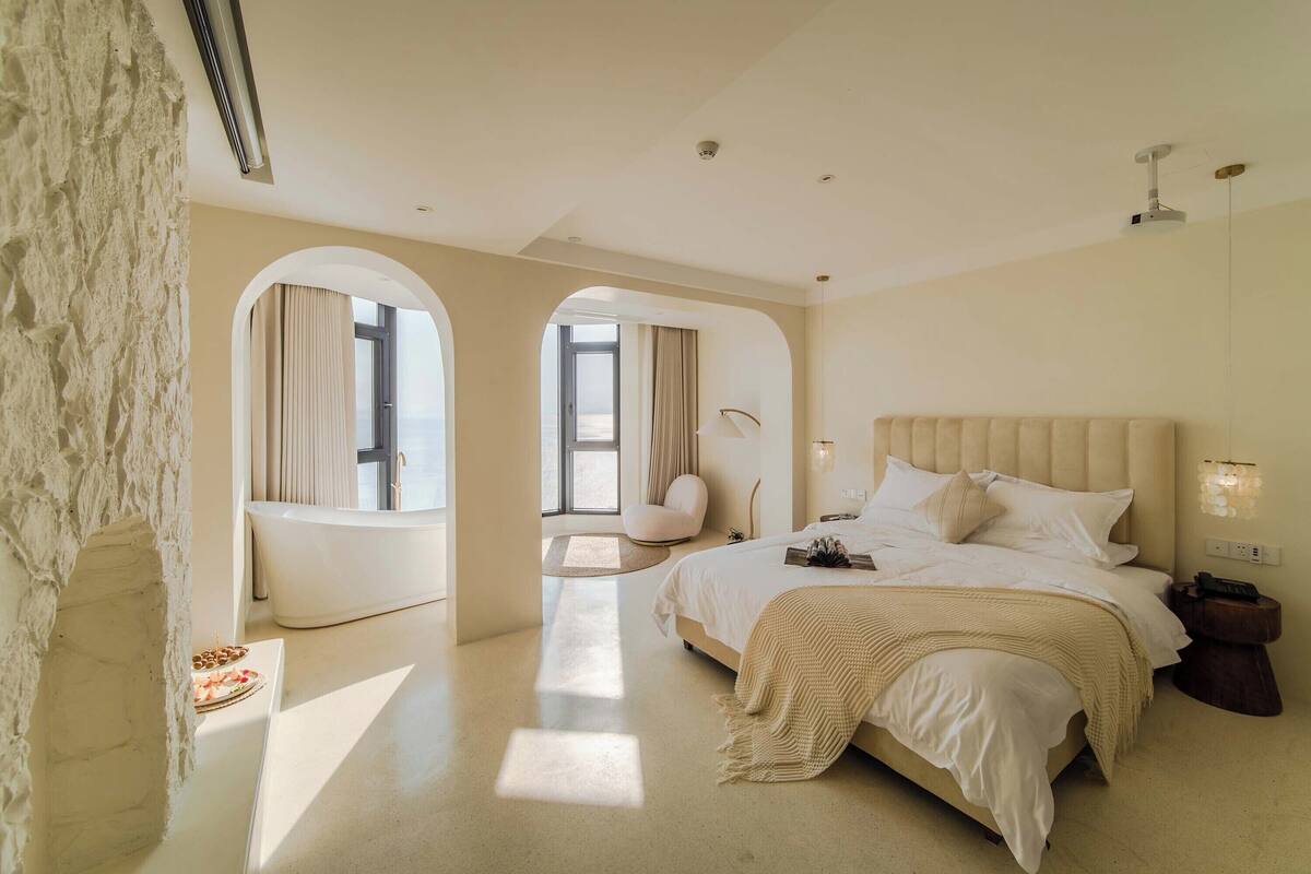 A modern bedroom with a large bed, a sitting area with a chair, and a view through an arched window.