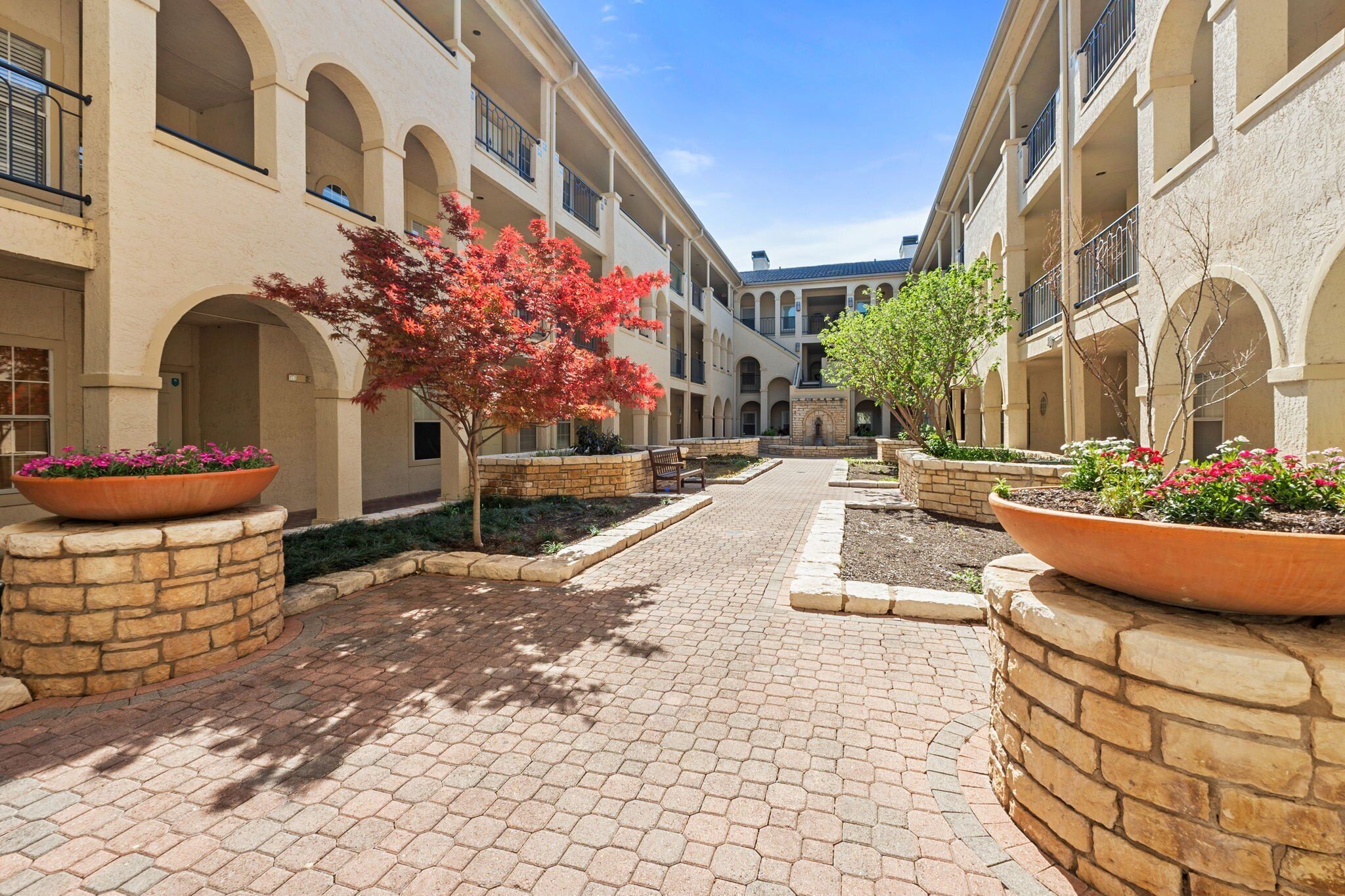 A courtyard with a paved walkway, stone planters with flowers, and a building with arched windows and balconies.