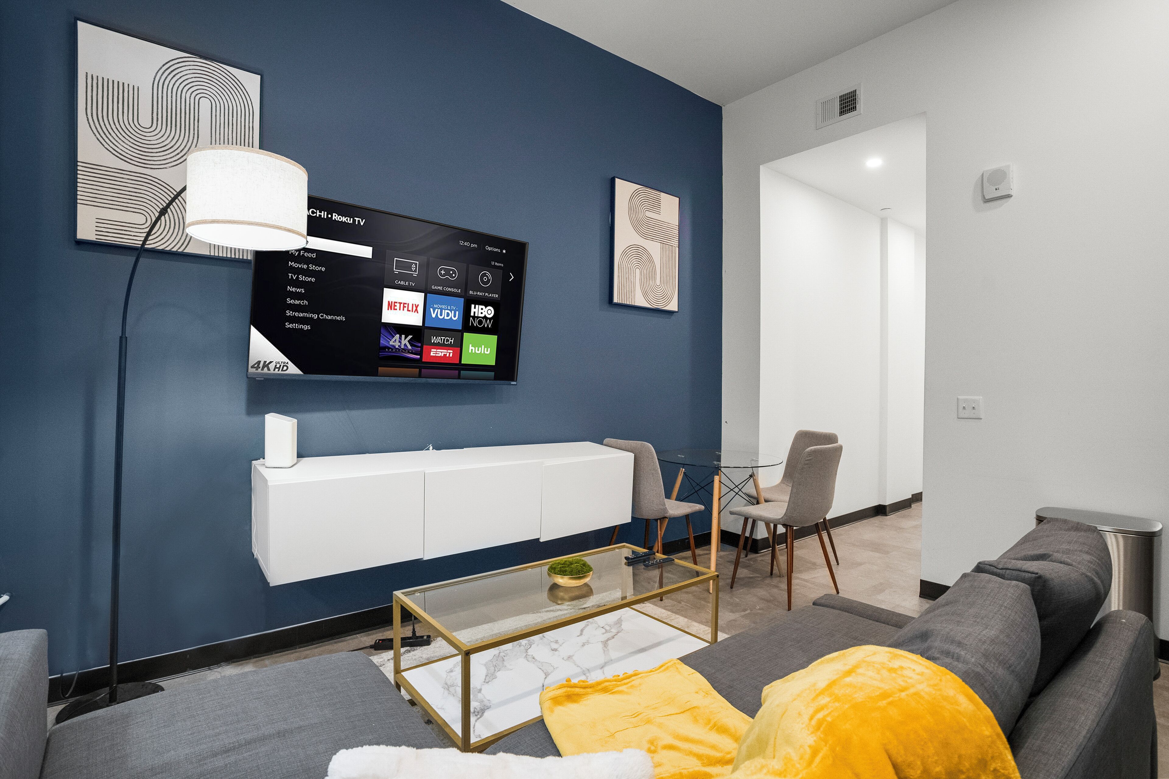 A modern living room with a mounted TV displaying streaming services, a glass coffee table, and a sofa with a yellow throw pillow.