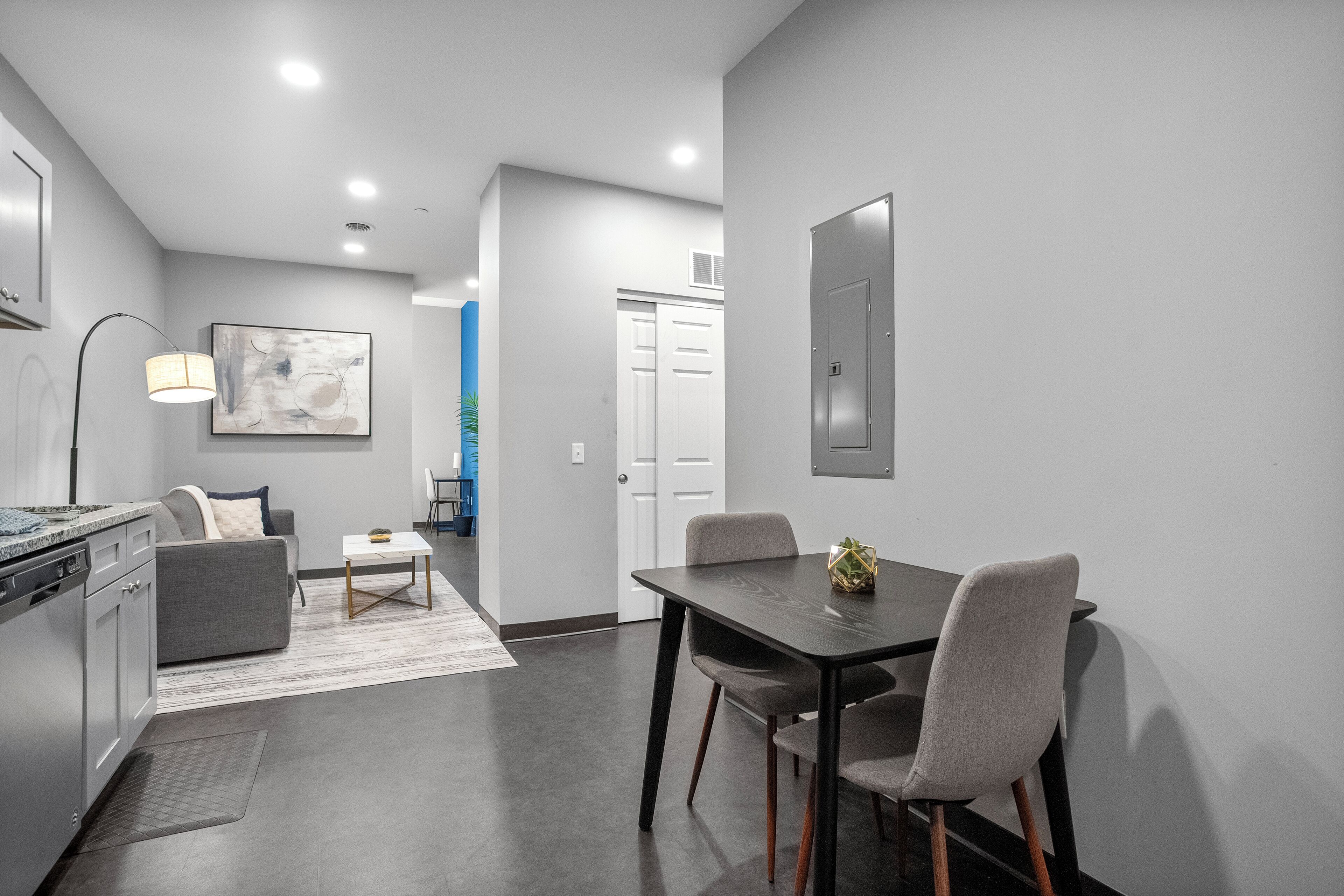 A modern kitchen with a built-in oven and dishwasher, a dining area with a dark wooden table and four chairs, and a living area with a gray sofa and a coffee table.