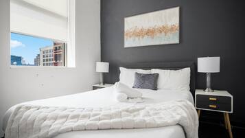 Habitación de hotel moderna con una cama grande, dos mesitas de noche con lámparas, vistas a la ciudad y una obra de arte minimalista en la pared.