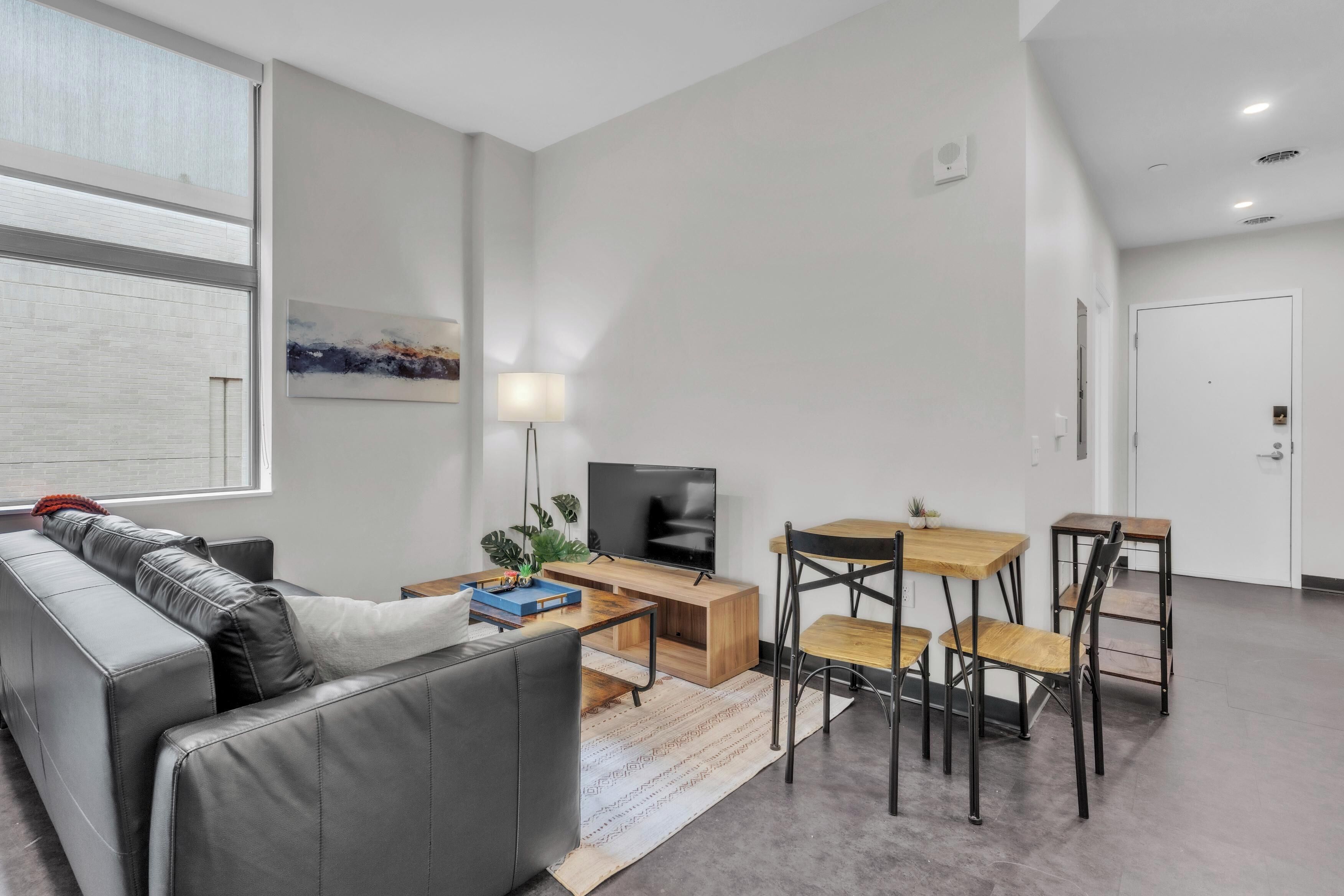 A modern living room with a grey sofa, a wooden coffee table, a TV, and a small dining area with chairs.