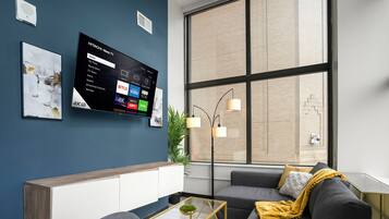 A modern living room with a mounted TV displaying streaming services, a sofa with cushions, a coffee table, and a large window with a view of a building.