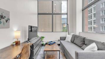 Una sala de estar moderna con un sofá, una mesa de centro de madera, un televisor y un amplio ventanal con vistas a edificios.