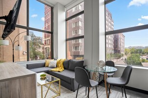 A modern living room with a gray sofa, a glass table, and a view of a cityscape through large windows.