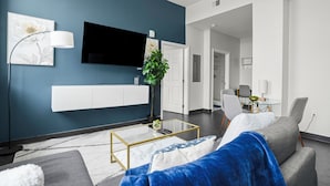 Ein modernes Wohnzimmer mit blauer Akzentwand, einem an der Wand montierten Fernseher, einem Glastisch und einem grauen Sofa.