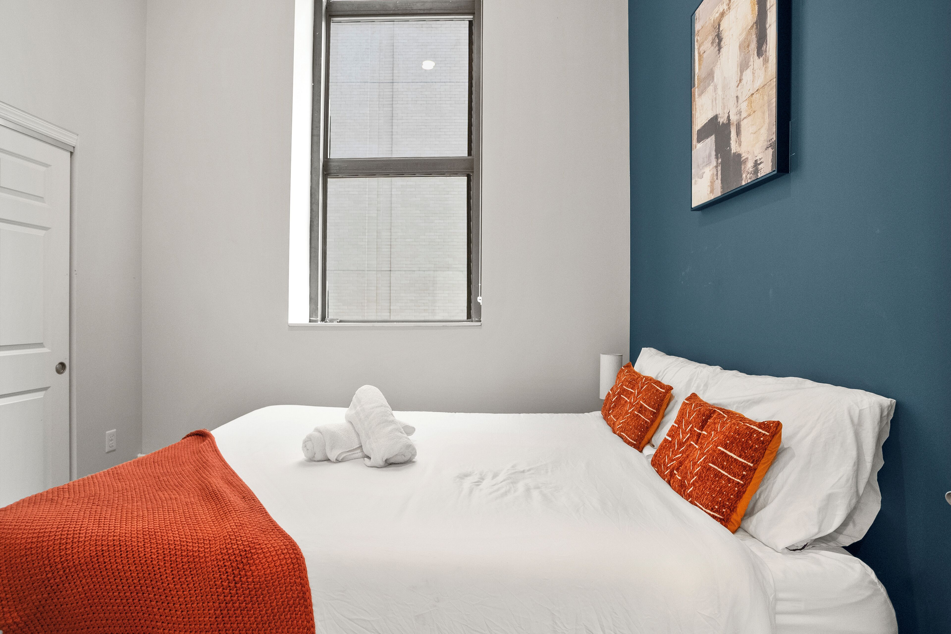 A neatly made bed with white linens and two orange pillows, a window with blinds, and a blue accent wall.