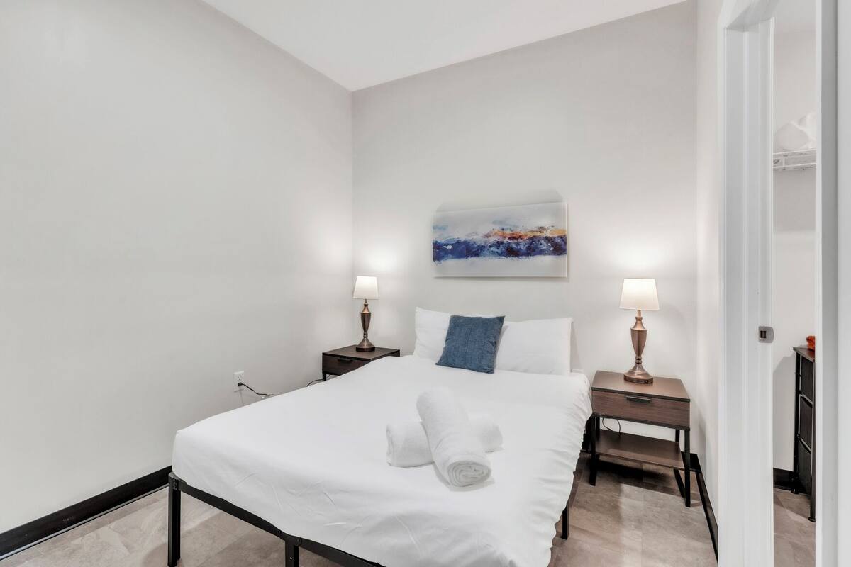 A hotel room with a bed, two bedside tables, a nightstand, a painting on the wall, and a door to the right.