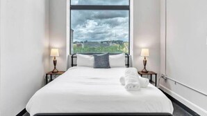 Ein Hotelzimmer mit großem Fenster, einem Bett mit weißer Bettwäsche, zwei Nachttischlampen und Blick auf die Stadt.