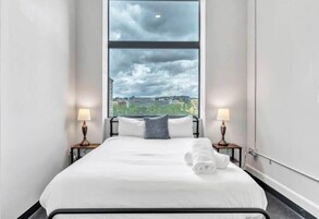 A hotel room with a large window, a bed with white linens, two bedside lamps, and a view of the cityscape.