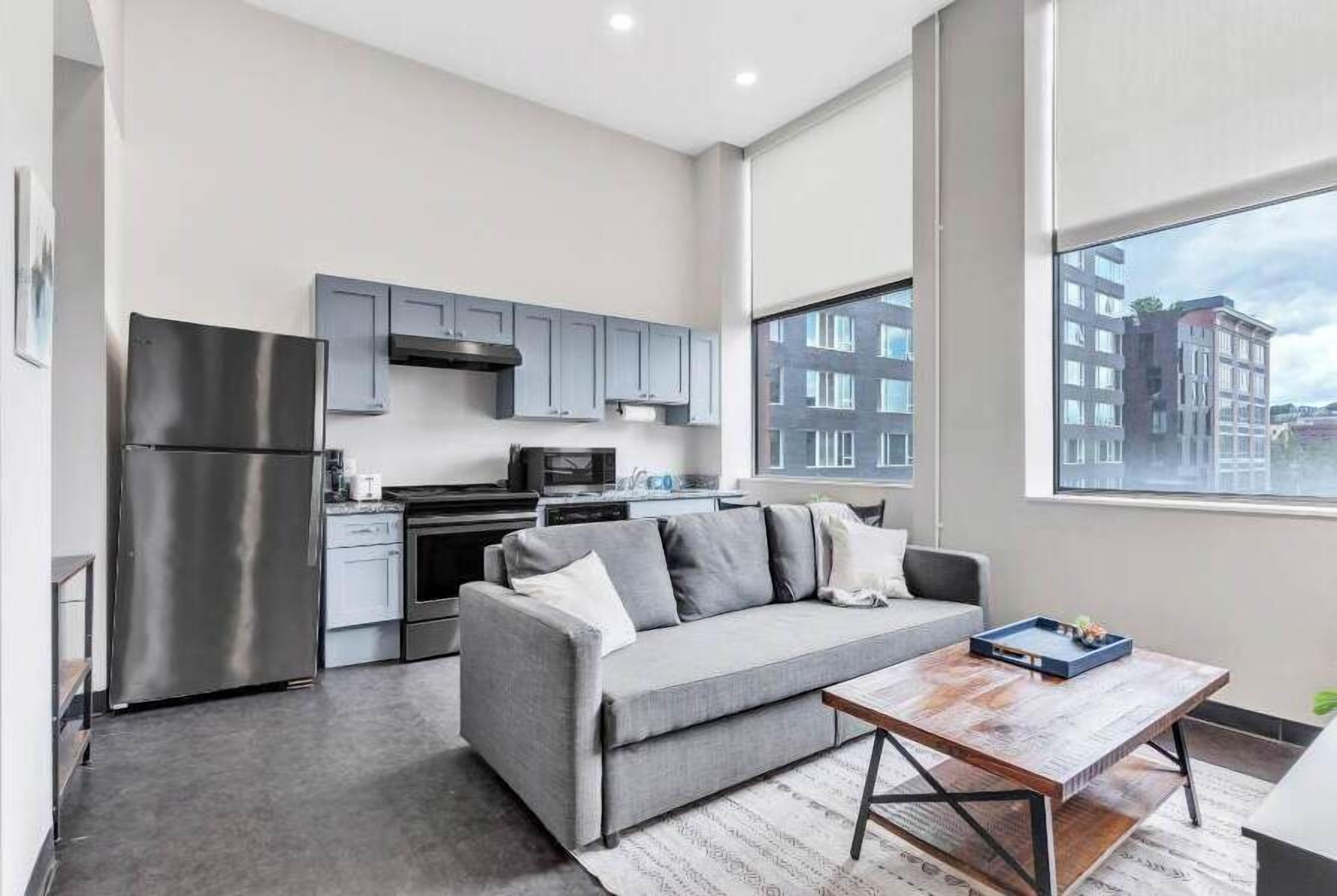 A modern kitchen with grey cabinets, a stainless steel refrigerator, and a grey sofa.