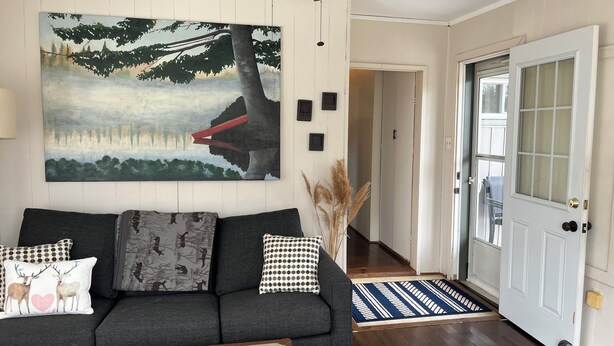 A living room with a gray sofa, a painting of a forest scene, a ceiling fan, and a door with a glass panel. - 5 min to the beach! Sun filled cottage in Turkey Point (Vittoria)
