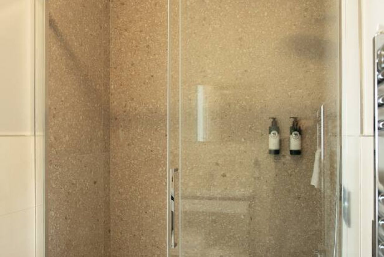 Ett duschutrymme med glasväggar, en beige kakelvägg och två flaskor schampo och balsam som står på väggen.