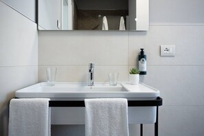 Ein modernes Badezimmer mit einem weißen Waschbecken, zwei Wasserhähnen und einem Spiegel darüber. Zwei Handtücher hängen an einem Haken, eine kleine Topfpflanze und eine Seife stehen auf dem Waschbecken.