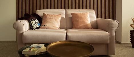 A beige sofa with two cushions, a small round table with a book, and a tall, narrow wooden panel wall.