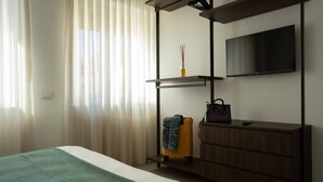 A hotel room with a bed, a TV mounted on the wall, a wooden dresser, a shelf with a vase, and a suitcase. - Palazzo Indaco - RESIDENZA RUBINO (Castel di Sangro)