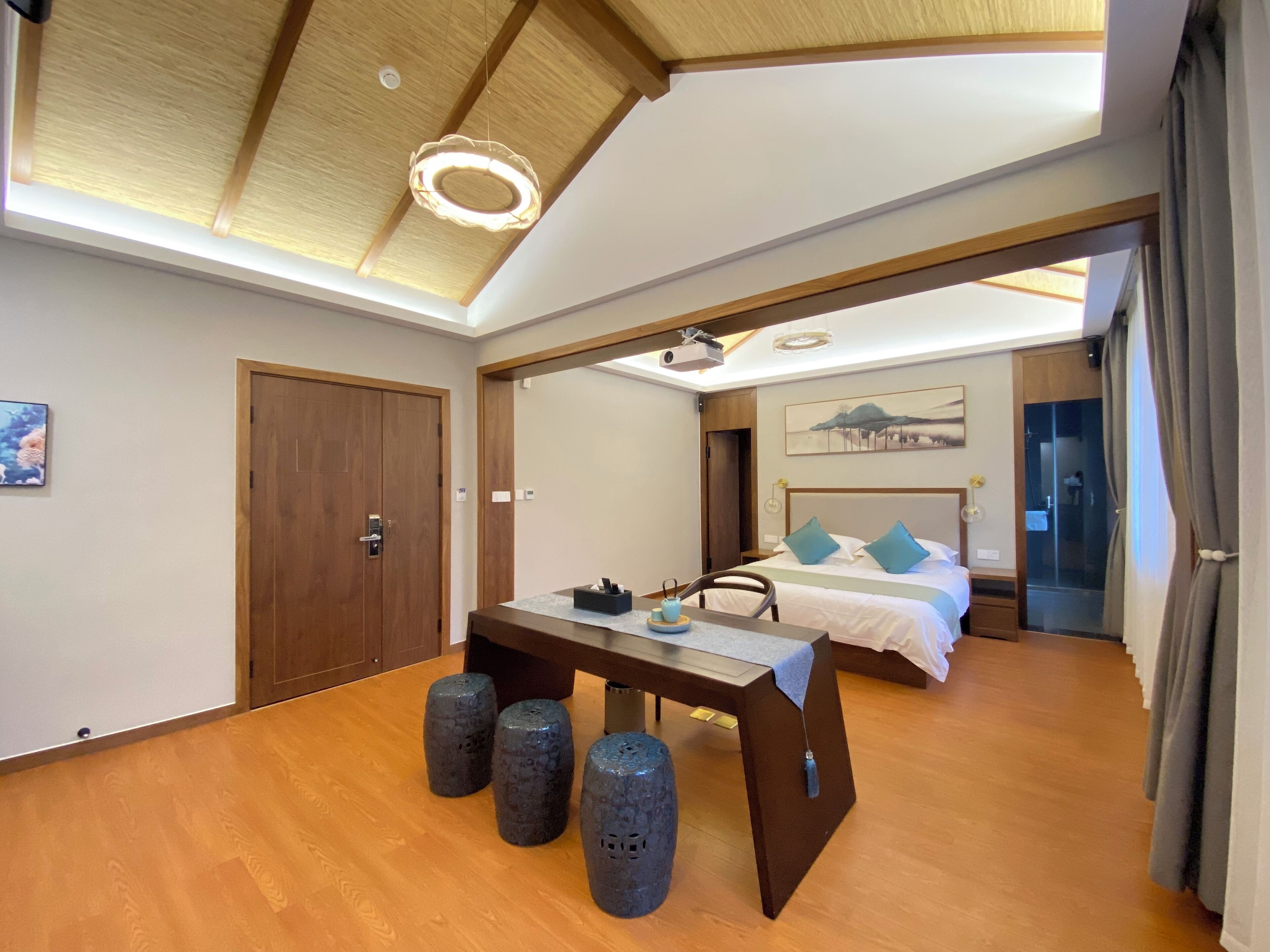 A modern hotel room with a wooden ceiling, a bed with blue pillows, a desk with a computer, and a large window.
