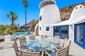 Un edificio blanco con detalles azules, una mesa de vidrio y sillas, rodeado por una piscina y palmeras.
