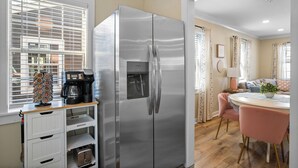 A modern kitchen with a stainless steel refrigerator, a coffee machine, and a dining area with a pink chair and a table. - Cottage Row: Daphne (Laurel)