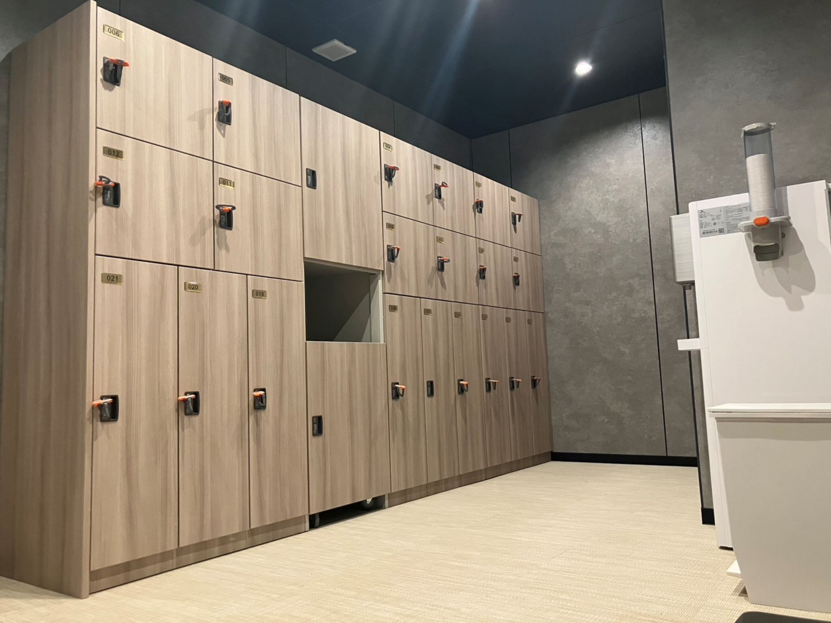 Une rangée de casiers en bois, chacun portant une étiquette numérotée, un réfrigérateur blanc et un mur gris.