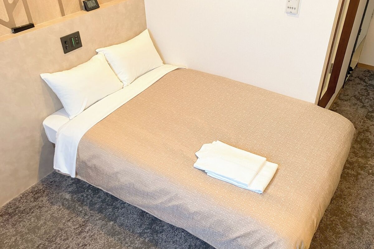 A neatly made bed with a beige bedspread and white pillows, a folded white towel on the bed, and a control panel on the wall.