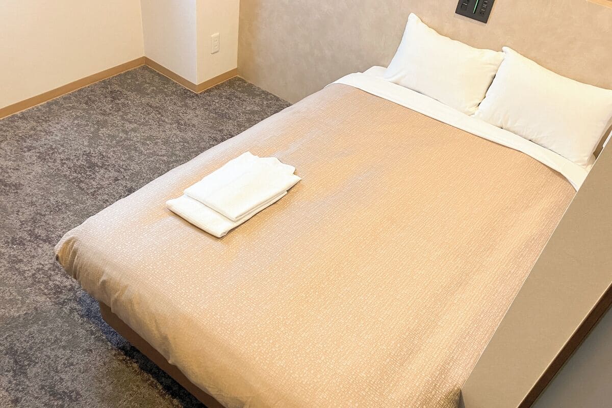 A neatly made bed with a beige bedspread and white pillows, a folded white towel on the bed, and a grey carpeted floor.
