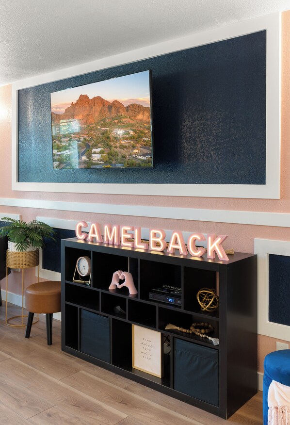 Una sala de estar moderna con un televisor montado que muestra un paisaje escénico, un mueble de entretenimiento negro con la palabra "CAMELBACK" iluminada y un cuadro enmarcado en la pared.