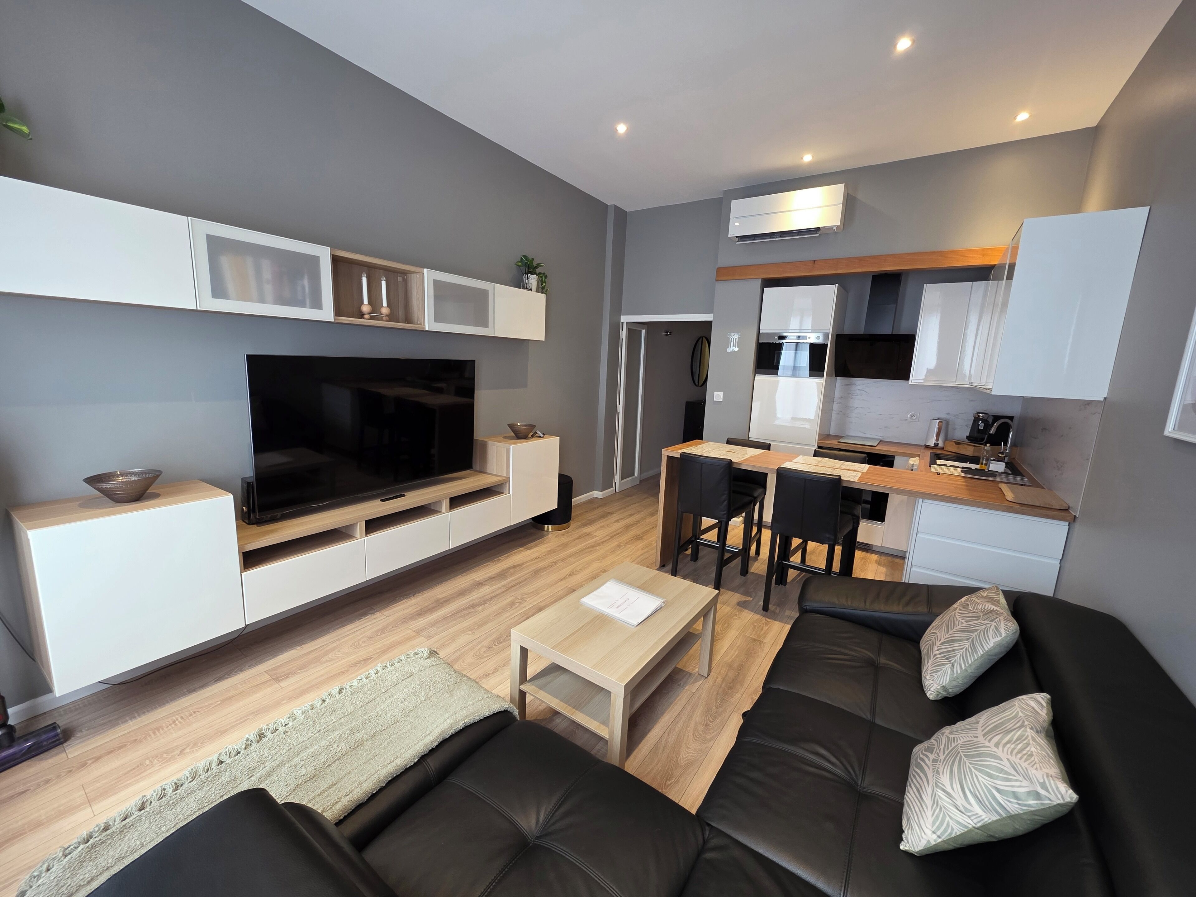 A modern living room with a flat-screen TV, a sofa, a coffee table, and a dining area with a kitchen in the background.