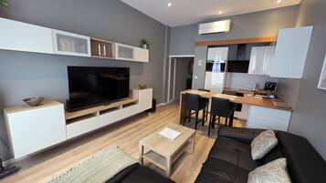 A modern living room with a flat-screen TV, a sofa, a coffee table, and a dining area with a kitchen in the background.