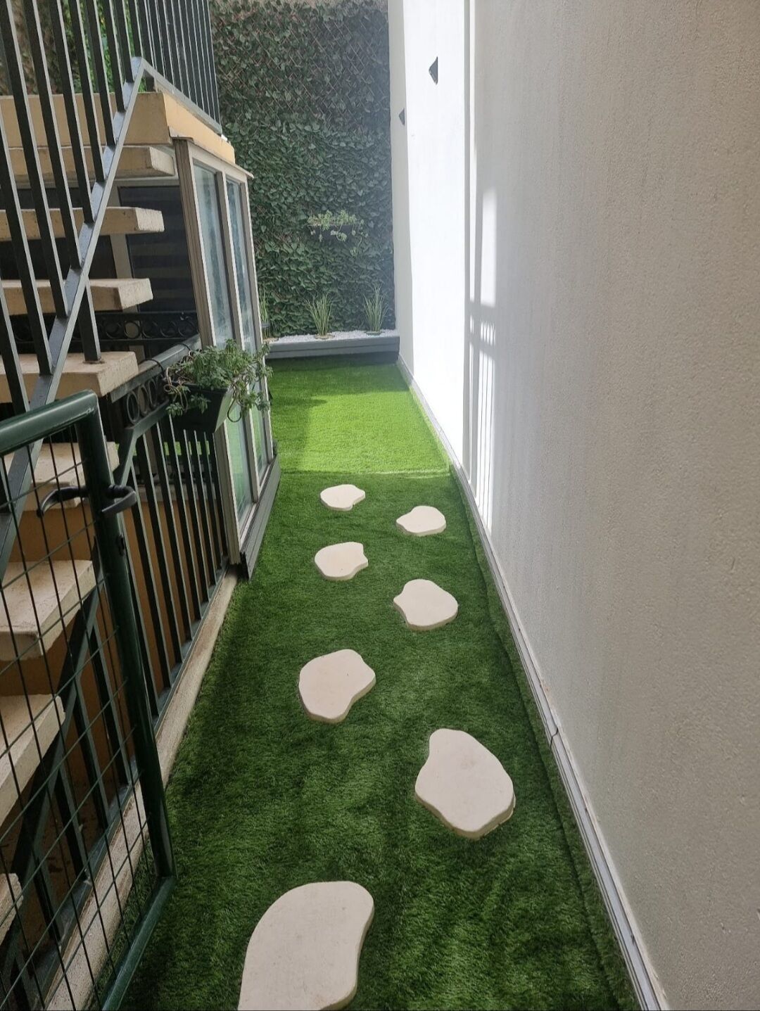 A narrow corridor with artificial green turf and stepping stones, bordered by a metal railing and a white wall.