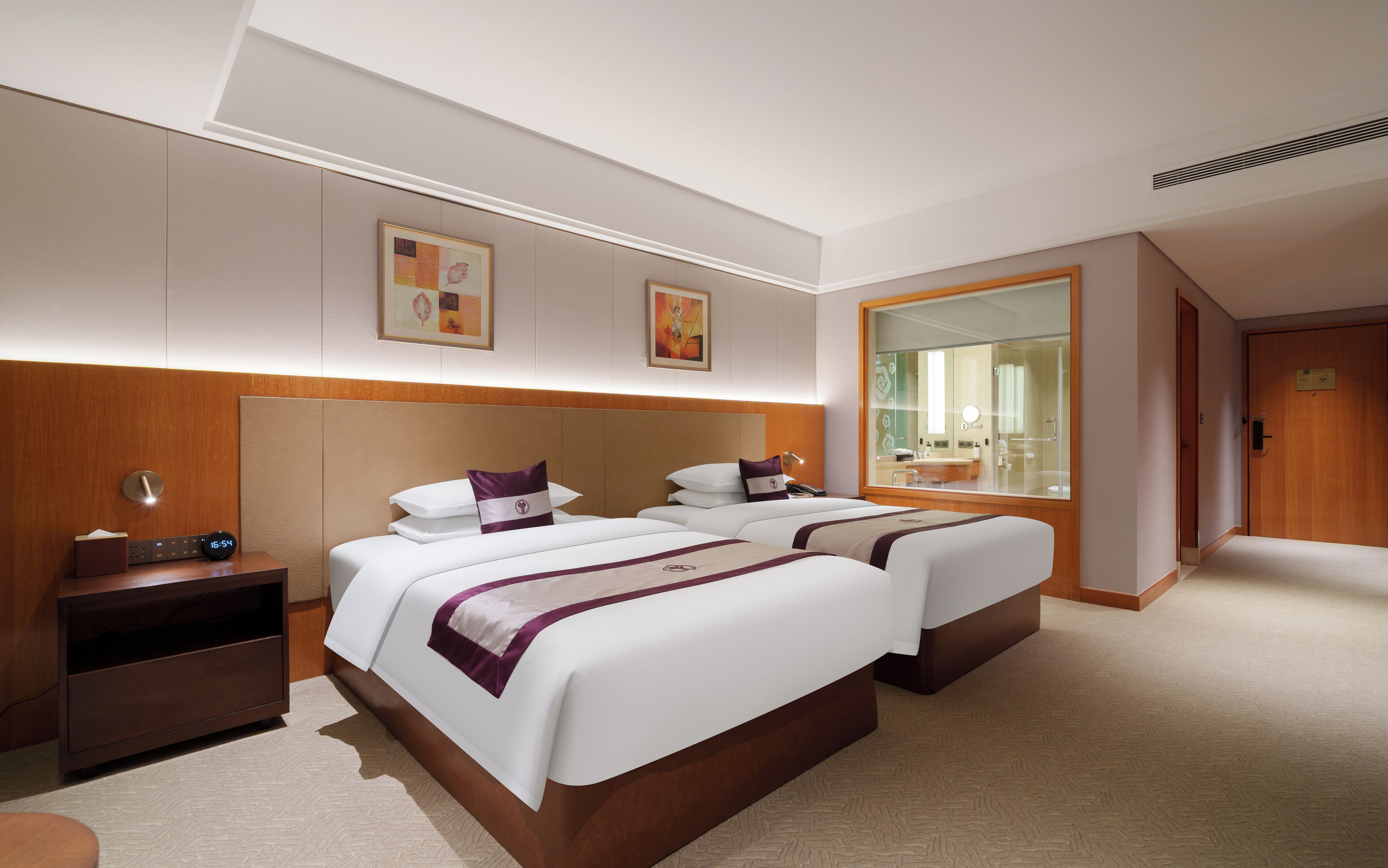 A hotel room with two beds, a bedside table with a clock, a mirror, and artwork on the walls.