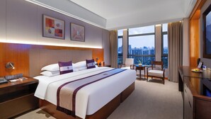 Une chambre d’hôtel avec un grand lit, un bureau, une chaise et une télévision. Elle comporte deux fenêtres avec des rideaux, et l’on peut admirer la vue sur la ville à travers ces fenêtres.