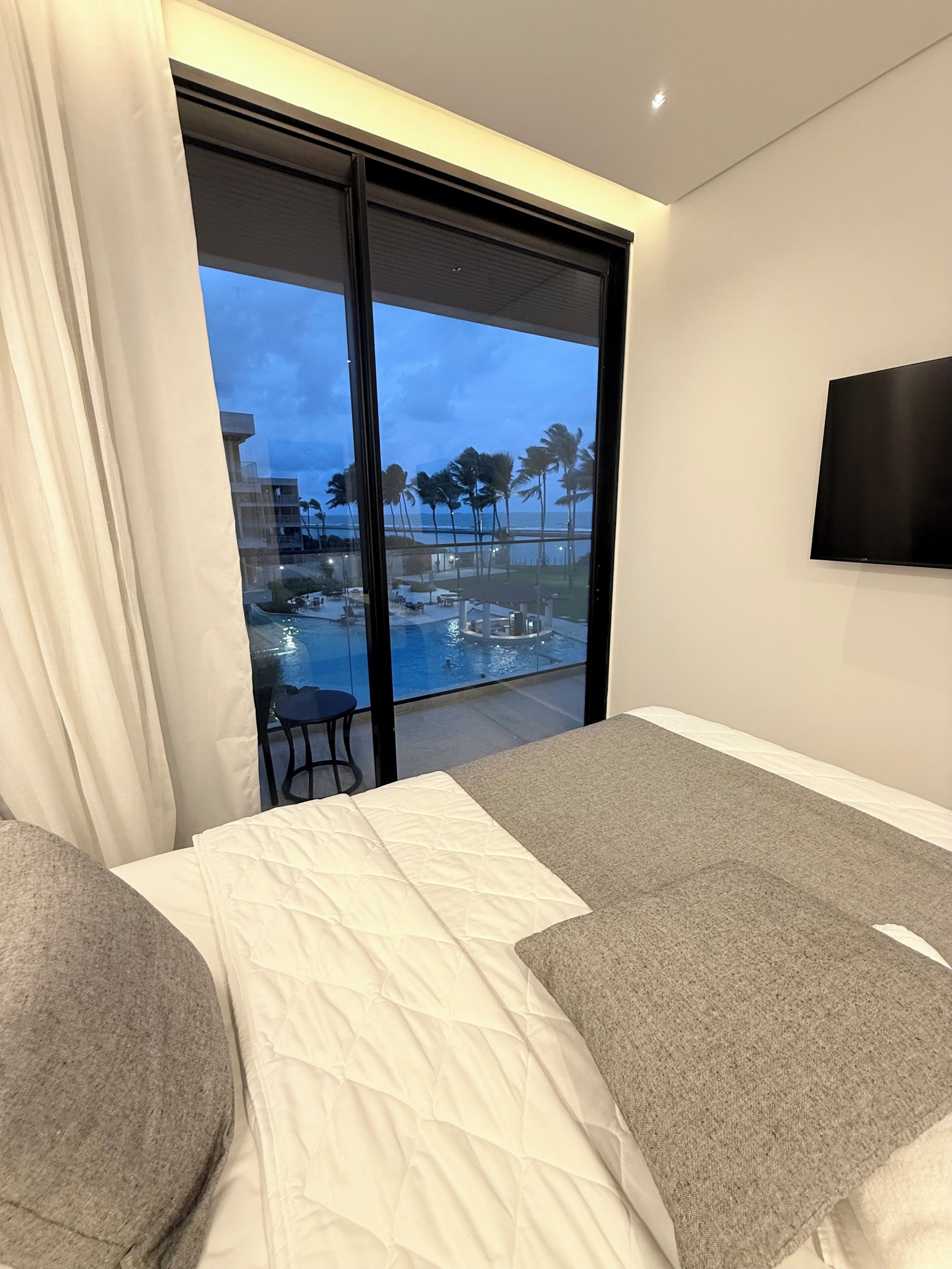 A hotel room with a large sliding glass door leading to a pool area, a flat-screen TV mounted on the wall, and a bed with a white quilted cover.