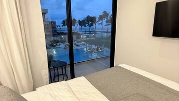 A hotel room with a large sliding glass door leading to a pool area, a flat-screen TV mounted on the wall, and a bed with a white quilted cover.