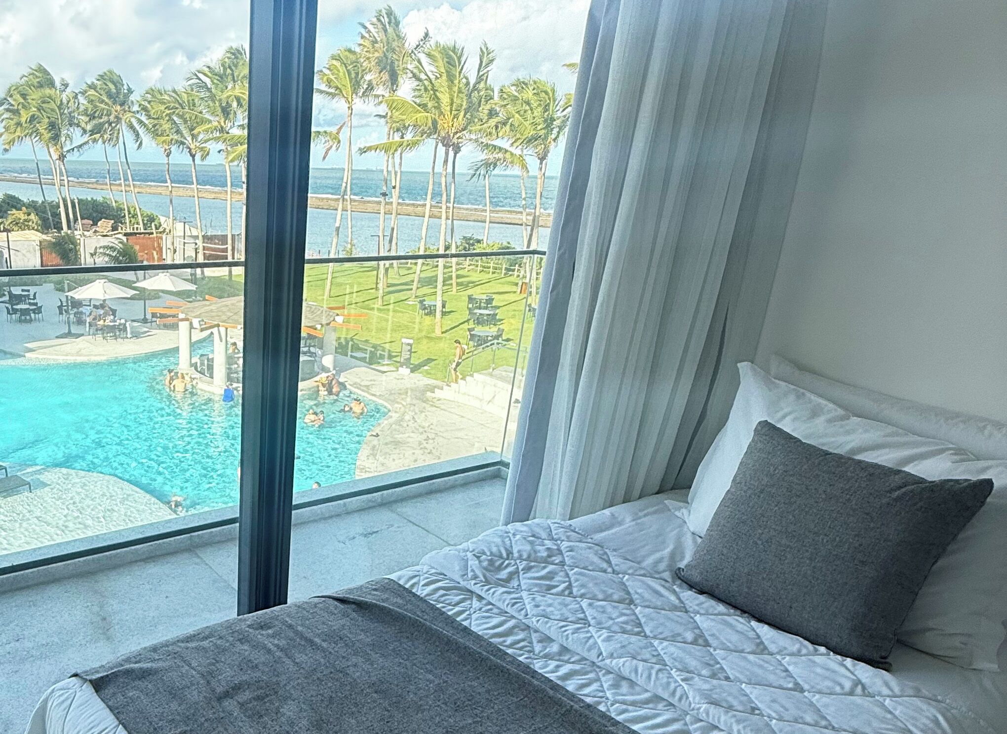 A hotel room with a bed, a view of a pool and palm trees, and a balcony with outdoor furniture.