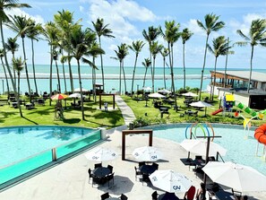 A resort pool area with multiple umbrellas, lounge chairs, and a view of the ocean. - Porto Alto Resort - Pé na Areia (Ipojuca)