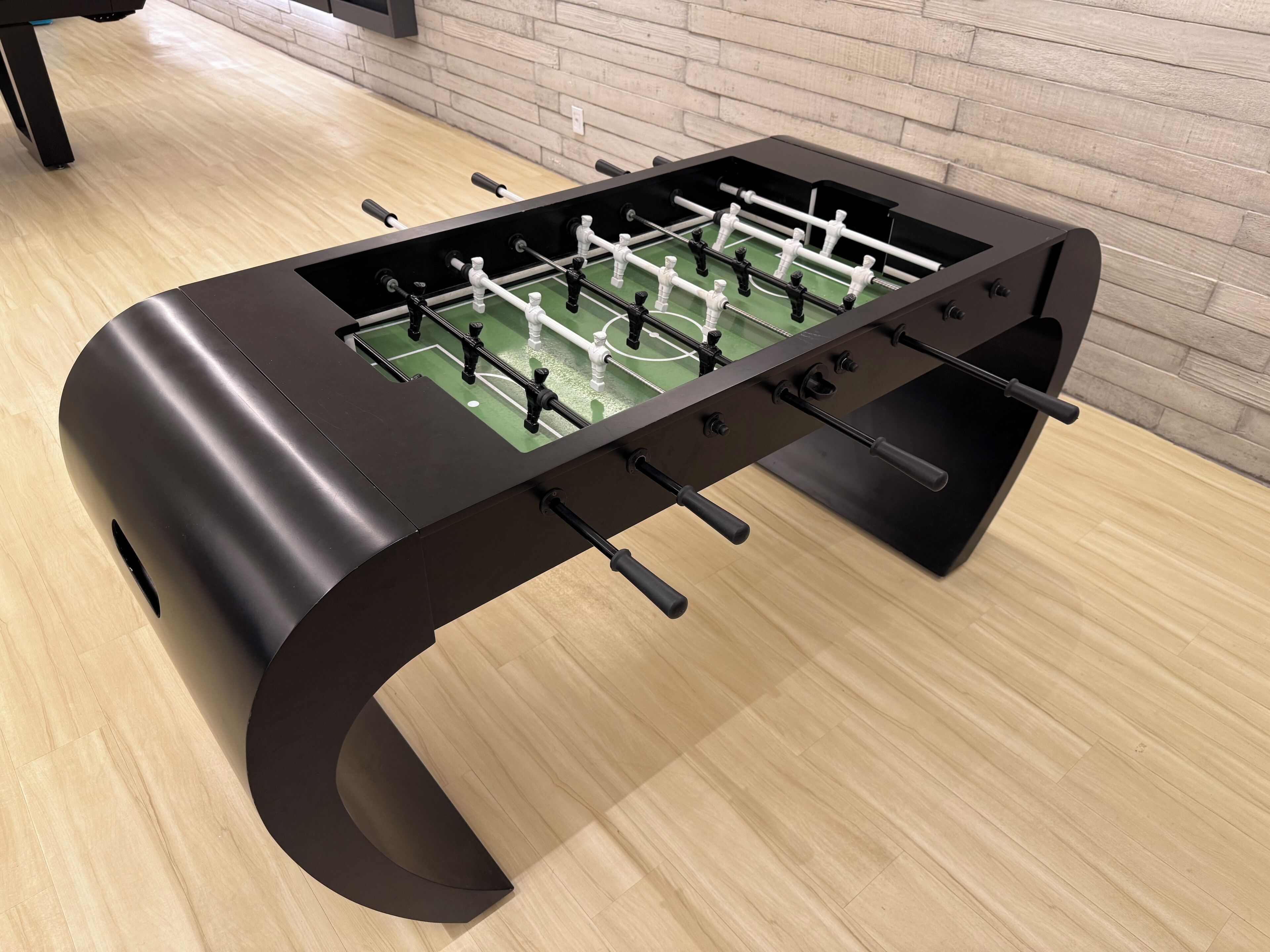 A modern foosball table with a curved design, located in a room with wooden flooring and a brick wall.