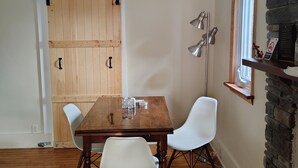 Comedor con mesa de madera y sillas blancas, riel de techo negro, chimenea de piedra y ventana con vistas.