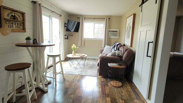 A cozy living area with a sofa, a coffee table, and a small dining table with stools. There is a TV mounted on the wall and a sliding door leading to another room.
